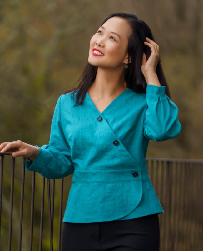 Woman wearing the Mauritius Top sewing pattern from Itch to Stitch on The Fold Line. A top pattern made in linen, linen blend, chambray, or broadcloth fabric, featuring a V-neck, wrap front, button closure, long sleeves with pleats, waistband, and peplum.