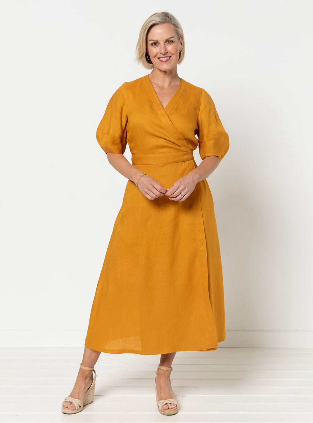 Woman wearing the Millicent Wrap Dress sewing pattern from Style Arc on The Fold Line. A dress pattern made in linen, cotton, silk, crepe, or rayon fabric, featuring a wrap bodice and skirt with a tie closure, V-neck, elbow length lantern sleeves, side pockets, and midi length.