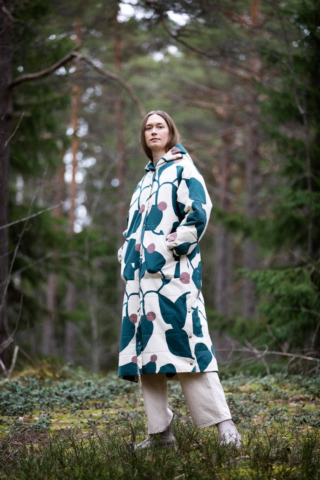 Woman wearing the Nietos Coat sewing pattern from TAUKO on The Fold Line. A quilted coat pattern made in cotton canvas fabric, featuring a front zipper, press studs, drawstring waist, side slits, raglan sleeves, pockets, and a hood.