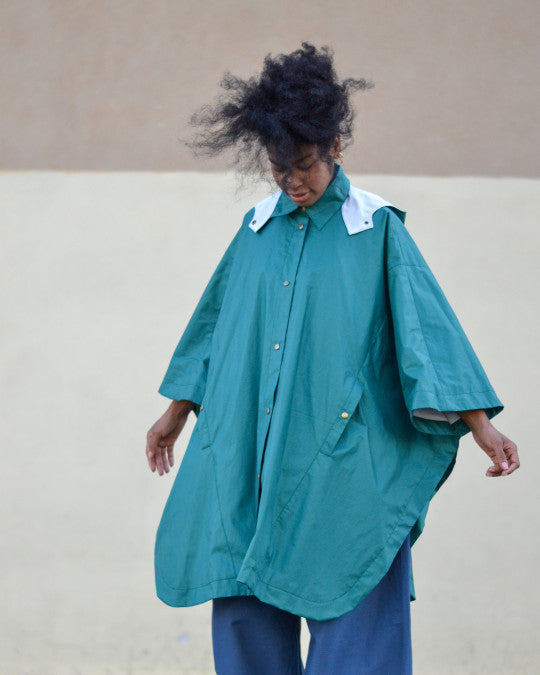Woman wearing the Oratge Cape sewing pattern from Pauline Alice on The Fold Line. A lined cape pattern made in gabardine, waxed cotton or linen, nylon, or wool fabric, featuring a collar, removable hood, snap buttons at the front and sides, welt pockets at the front, and patch pockets inside.