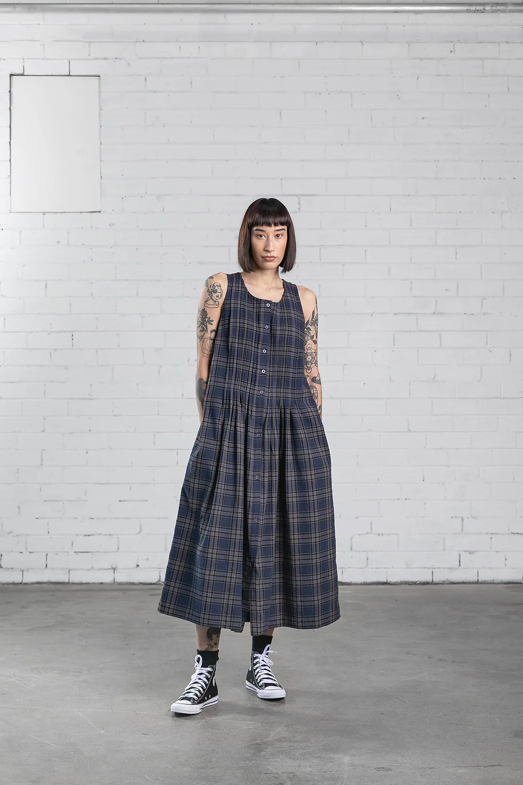 Woman wearing the Page Dress sewing pattern from Elbe Textiles on The Fold Line. A sleeveless dress pattern made in cotton, linen, silk noil, hemp, or linen viscose fabric, featuring a rounded neckline, button front, tucks running down the front and back bodice that open out into a pleated skirt, in-seam pockets, side splits, and midi length.
