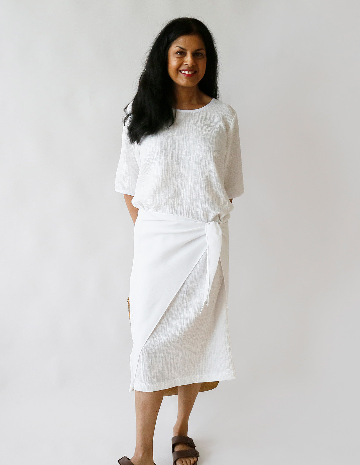 Woman wearing the Pareo Dress sewing pattern from The Maker's Atelier on The Fold Line. A pull-on dress pattern made in cotton, linen, silk, Tana lawn, tencel, viscose, or poly/viscose mix fabric, featuring a round neckline, short sleeves, integrated wrap ties, and midi length.