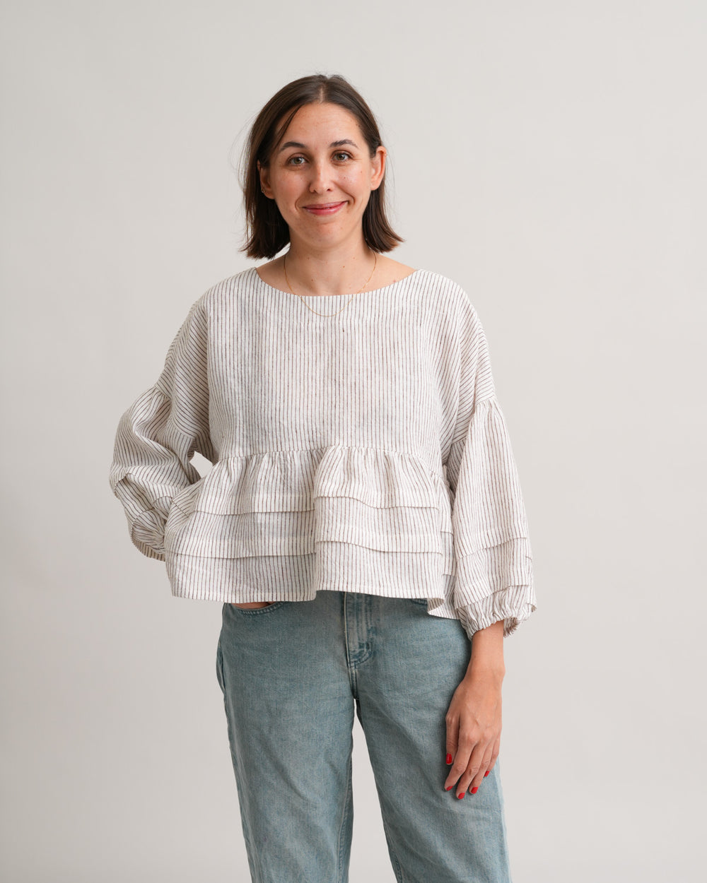 Woman wearing the Pintuck Poet Blouse sewing pattern from Matchy Matchy Sewing Club on The Fold Line. A top pattern made in cotton or linen fabric, featuring a relaxed boxy silhouette, rounded neckline, long puff sleeves with pintucks, gathered peplum with pintucks, and bow closures at the back.