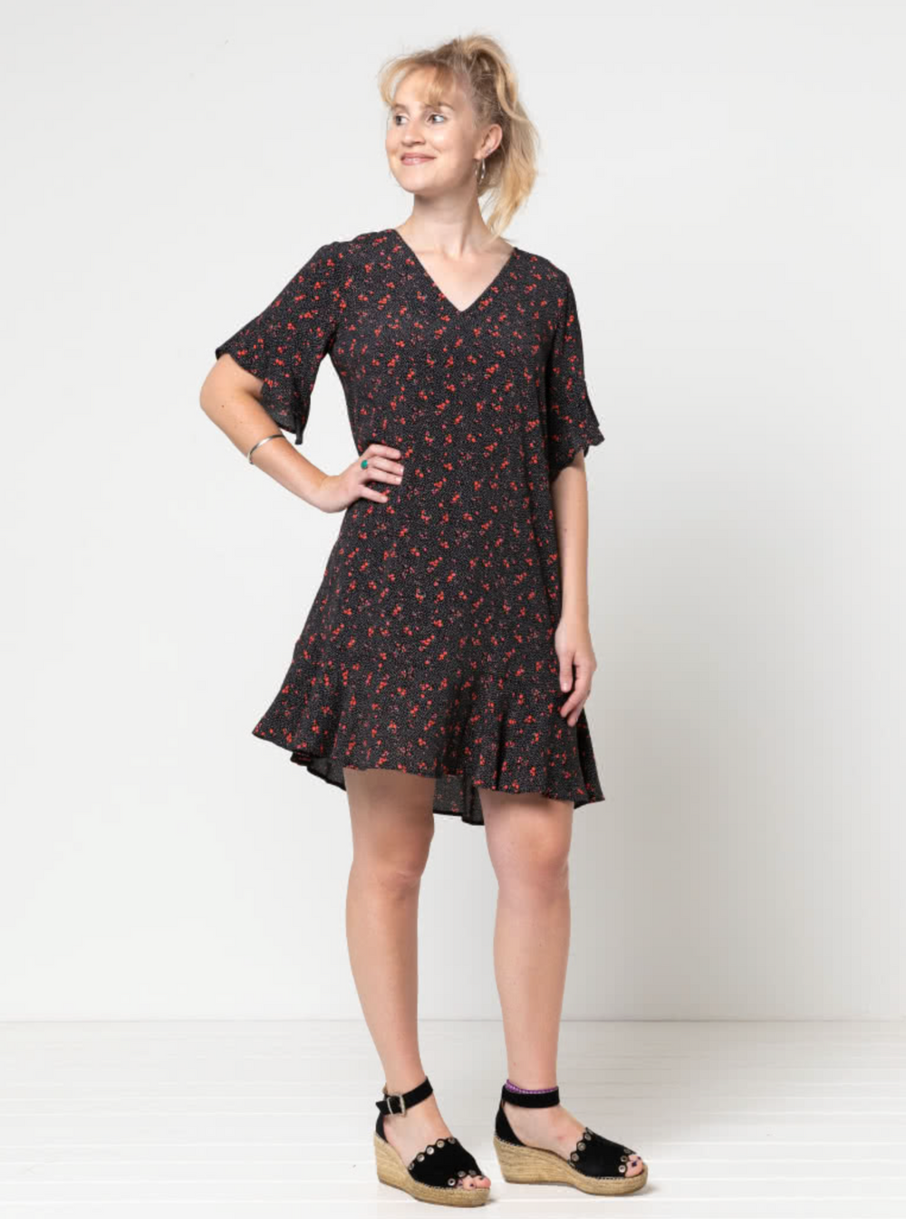 Woman wearing the Pixie Woven Dress sewing pattern from Style Arc on The Fold Line. A knee length dress pattern made in rayon, crepe, or silk fabric, featuring a slight A-line shape, V-neck, bust darts, short sleeves with flounces and a hem flounce.
