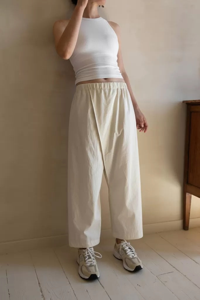 Woman wearing the Pleat Asymmetrical Pants sewing pattern from Ready to Sew on The Fold Line. A pants pattern made in cotton poplin, cotton sateen, or chambray fabric, featuring an elastic waist, front pleat, straight leg, and 7/8 length.