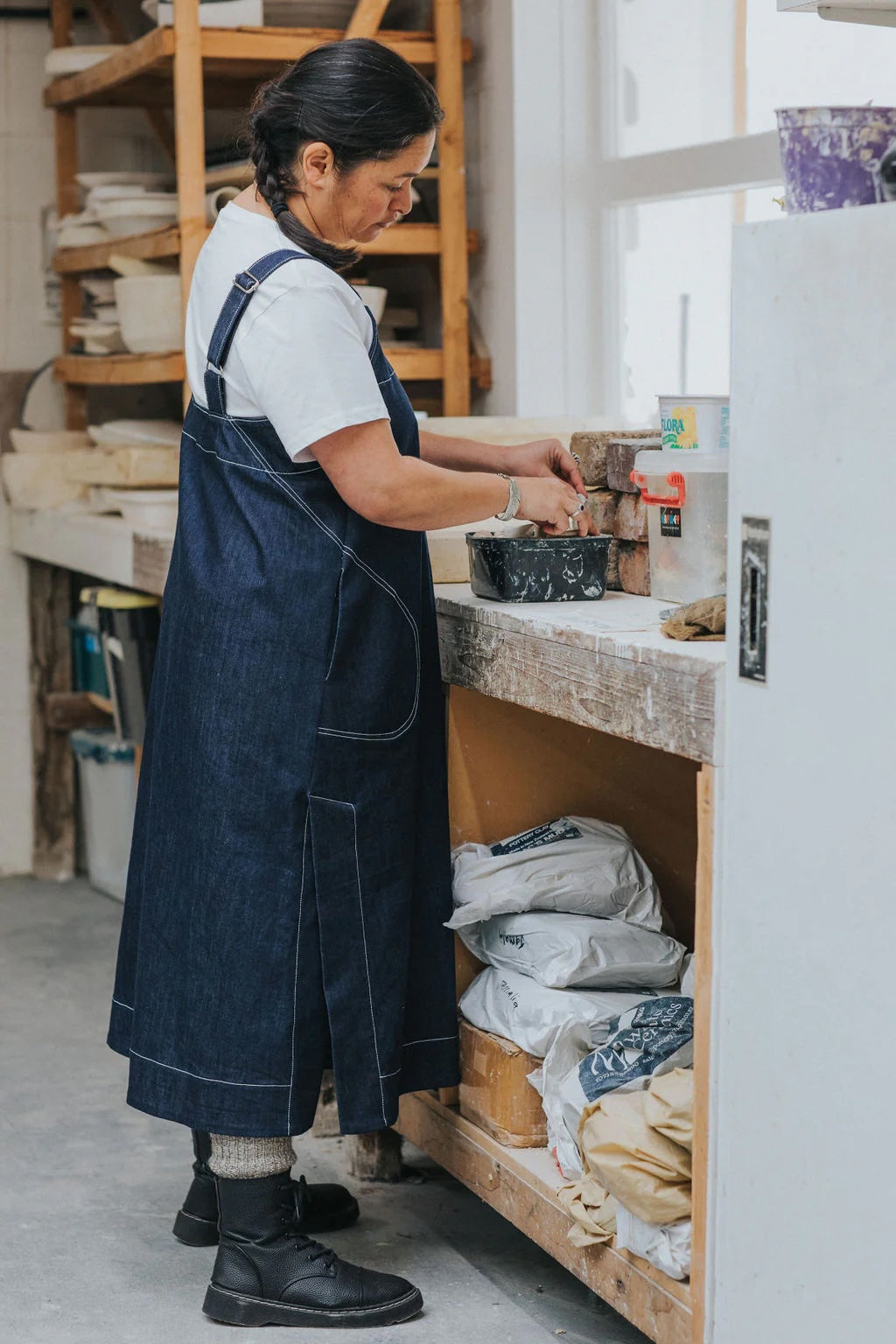 Woman wearing the Ray Pinafore sewing pattern from Paper Theory Patterns on The Fold Line. A dress pattern made in cotton, linen, or denim fabric, featuring a trapeze shape, dungaree straps, side seam pockets, a wide hem with side splits, and midi length.