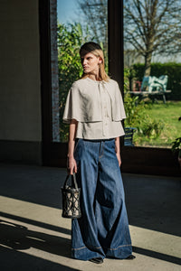 Woman wearing the Rima Blouse sewing pattern from Fibre Mood on The Fold Line. A sleeveless blouse pattern made in viscose (crepe), lyocell, satin, chambray, fine corduroy, denim, leather, seersucker, voile, muslin, crepon, poplin, raw silk, nylon, or organza fabric, featuring a round neckline with a tie, button front, and cape.