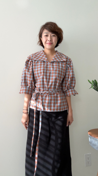 Woman wearing the Roma Top sewing pattern from Sewing Therapy on The Fold Line. A blouse pattern made in cotton lawn, voile, Swiss dot, linen or linen blends, tencel, cupro, rayon, double gauze, poplin, cotton sateen, chambray, seersucker, lightweight denim or canvas, viscose, silk crepe, or chiffon fabric, featuring an oversized silhouette, V-neckline, collar, drop shoulders, pleated mid-length sleeves, and waist ties.
