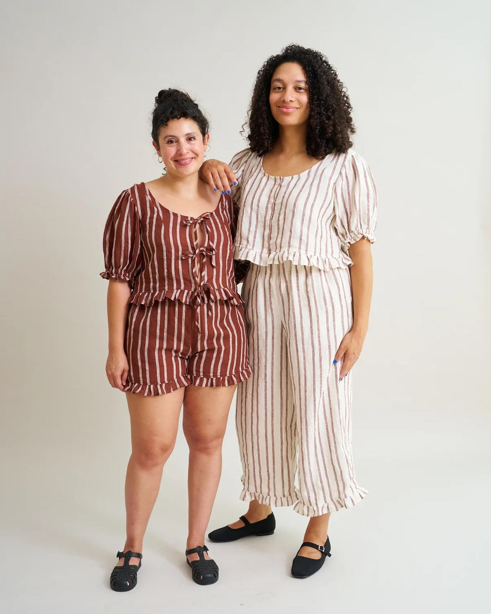 Women wearing the Ruffle Brunch Set sewing pattern from Matchy Matchy Sewing Club on The Fold Line. A co-ord pattern made in linen, cotton, or rayon fabric, featuring a cropped top with a wide scoop neckline, princess seams, short puff sleeves, tie or button front, and hem ruffles, and cropped pants or shorts with an elastic waist, pockets, and hem ruffles.
