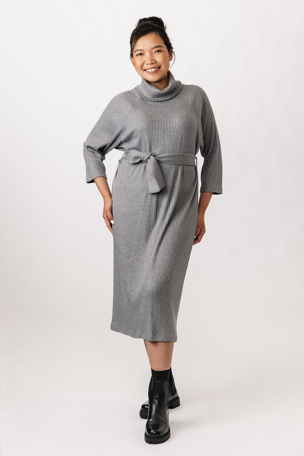 Woman wearing the Salla Sweater Dress sewing pattern from Named on The Fold Line. A dress pattern made in knit fabric, featuring a straight cut, chunky turtleneck, wrist-length raglan sleeves, a wide belt, and midi length.