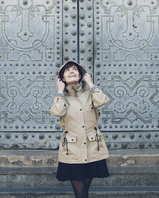 Woman wearing the Serra Jacket sewing pattern from Pauline Alice on The Fold Line. A raincoat pattern made in gabardine, linen, cotton twill, nylon, ripstop, or wool fabric, featuring a full lining, raglan sleeves, storm shield, stand-up collar, zip closure and button placket, drawstring waist, three flap pockets, detachable hood, and mid-thigh length. 