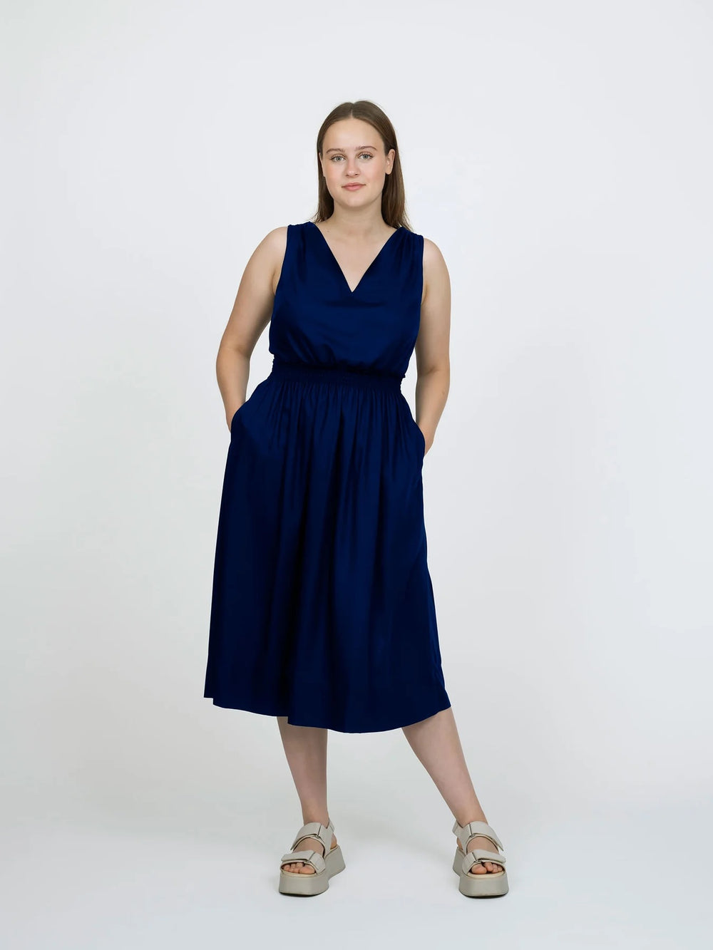 Woman wearing the Smocked Dress sewing pattern from The Assembly Line on The Fold Line. A sleeveless dress pattern made in lightweight fabric, featuring a V-neckline, elastic shirring at the shoulders and waist, side seam pockets, and midi length.