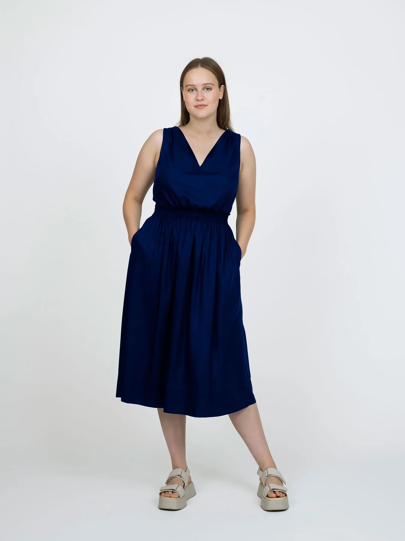 Woman wearing the Smocked Dress sewing pattern from The Assembly Line on The Fold Line. A sleeveless dress pattern made in lightweight fabric, featuring a V-neckline, elastic shirring at the shoulders and waist, side seam pockets, and midi length.