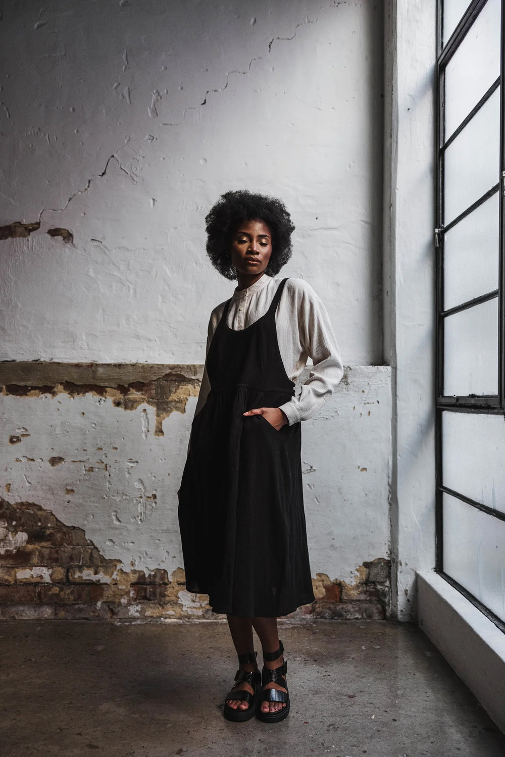 Woman wearing the Steele Pinafore sewing pattern from Elbe Textiles on The Fold Line. A dress pattern made in tencel, rayon, washed linen, silk noil, or soft cotton fabric, featuring adjustable straps, button and loop side closures, pockets, and below knee length.