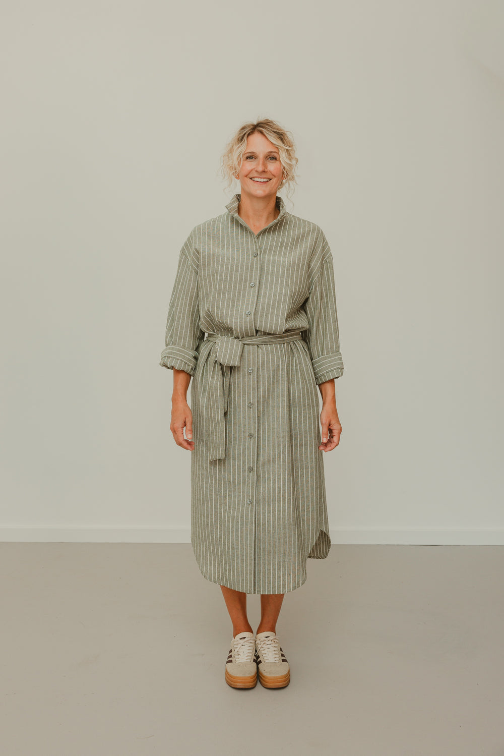 Woman wearing the Studio Dress sewing pattern from The Avid Seamstress on The Fold Line. A shirt dress pattern made in linen, cotton, cotton needlecord, poplin, lawn, shirting, viscose, or bamboo fabric, featuring a relaxed fit, collar, dropped shoulders, three-quarter sleeves with cuffs, button front, tie belt, side pockets, curved hem, and midi length.