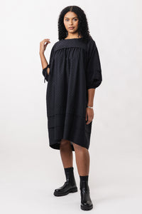 Woman wearing the Syli Dress sewing pattern from Named on The Fold Line. A dress pattern made in cotton, linen, tencel, or rayon fabric, featuring generous gathering, a bound neckline, pleat details, side pockets, mid-length sleeves, and knee length.