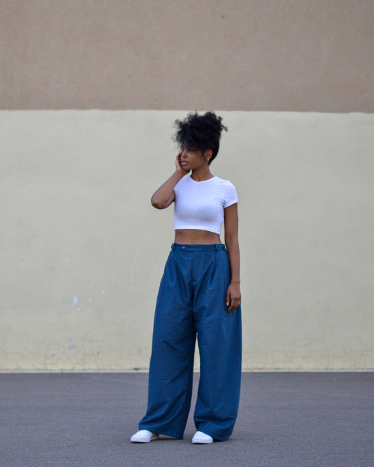 Woman wearing the Tardor Trousers sewing pattern from Pauline Alice on The Fold Line. A trousers pattern made in cotton or wool gabardine, poplin, chambray, linen, flannel, corduroy, or crepe fabric, featuring a high waist, wide legs with no side seams, front pleats, fly front, fitted waistband with belt loops, and adjustable pleat detail on the sides. 