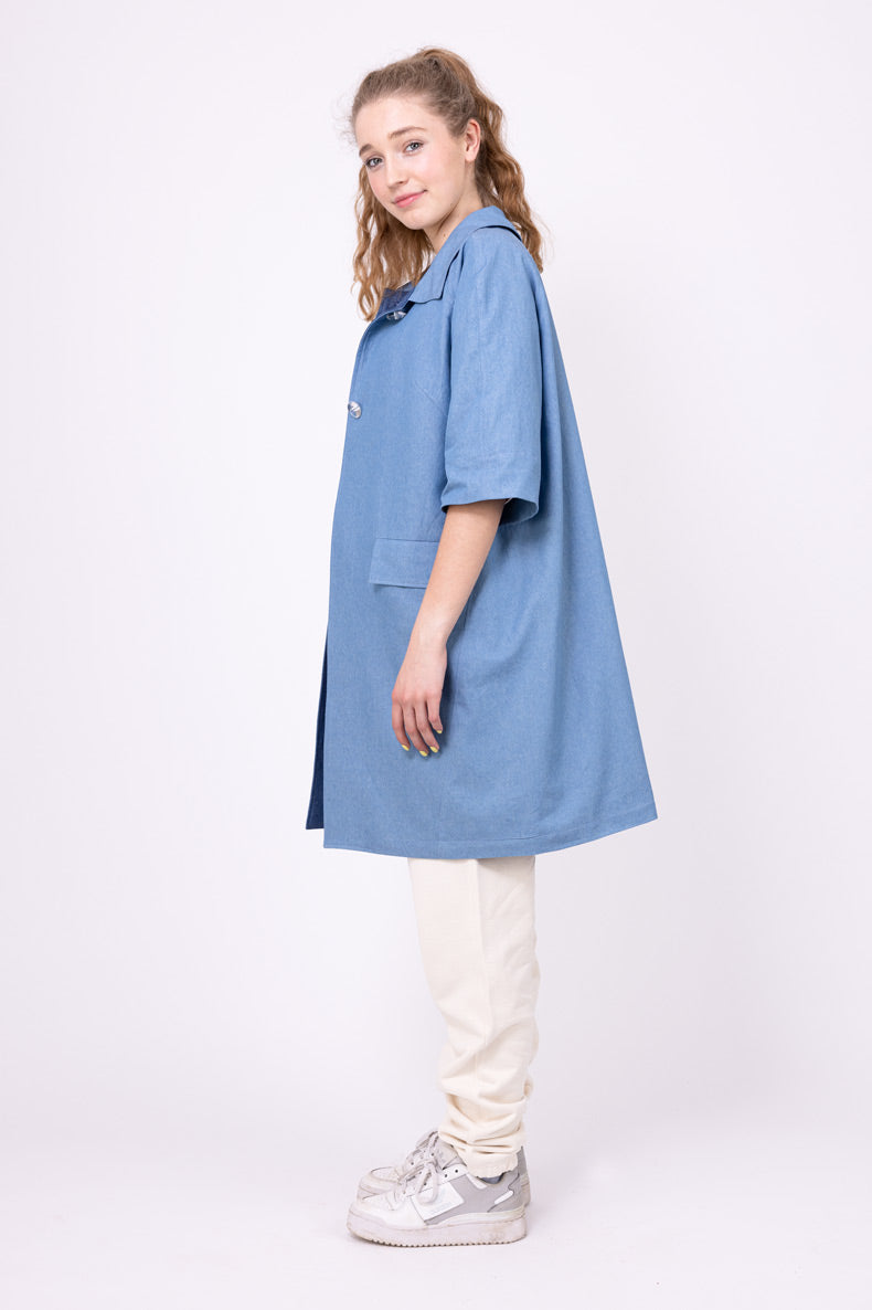 Woman wearing the Tessa Coat sewing pattern from Schnittchen Patterns on The Fold Line. A knee-length coat pattern made in denim, cotton, linen, or light wool fabric, featuring a collar, mid-length sleeves, pockets with flaps, bound buttonholes, and a lining.