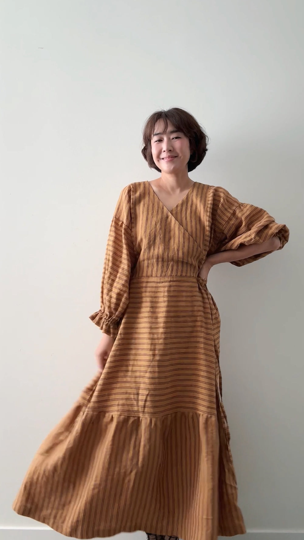 Woman wearing the Tie Dress No. 2 sewing pattern from Sewing Therapy on The Fold Line. A dress pattern made in linen or linen blends, cotton lawn, voile, rayon, viscose, tencel (lyocell), double gauze, or lightweight flannel fabric, featuring a wrap front bodice with a V-neck, long sleeves with elasticised ruffle cuffs, and a tiered skirt.