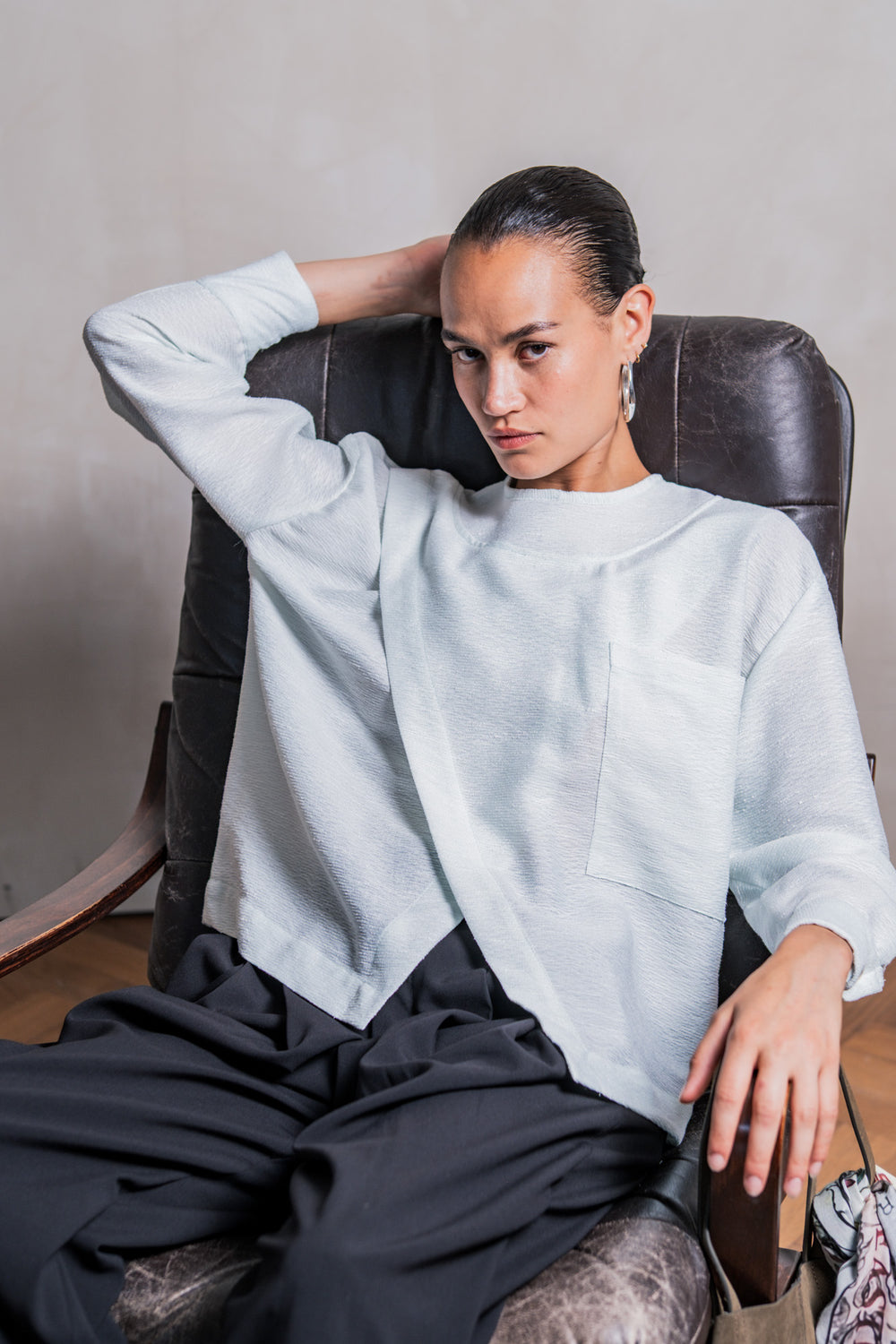 Woman wearing the Veya Blouse sewing pattern from Fibre Mood on The Fold Line. A top pattern made in crepe, viscose, poplin, lyocell, chambray, linen, seersucker, or broderie anglaise fabric, featuring a loose fit, round neck, asymmetrical overlap, pocket, and long sleeves with cuffs.