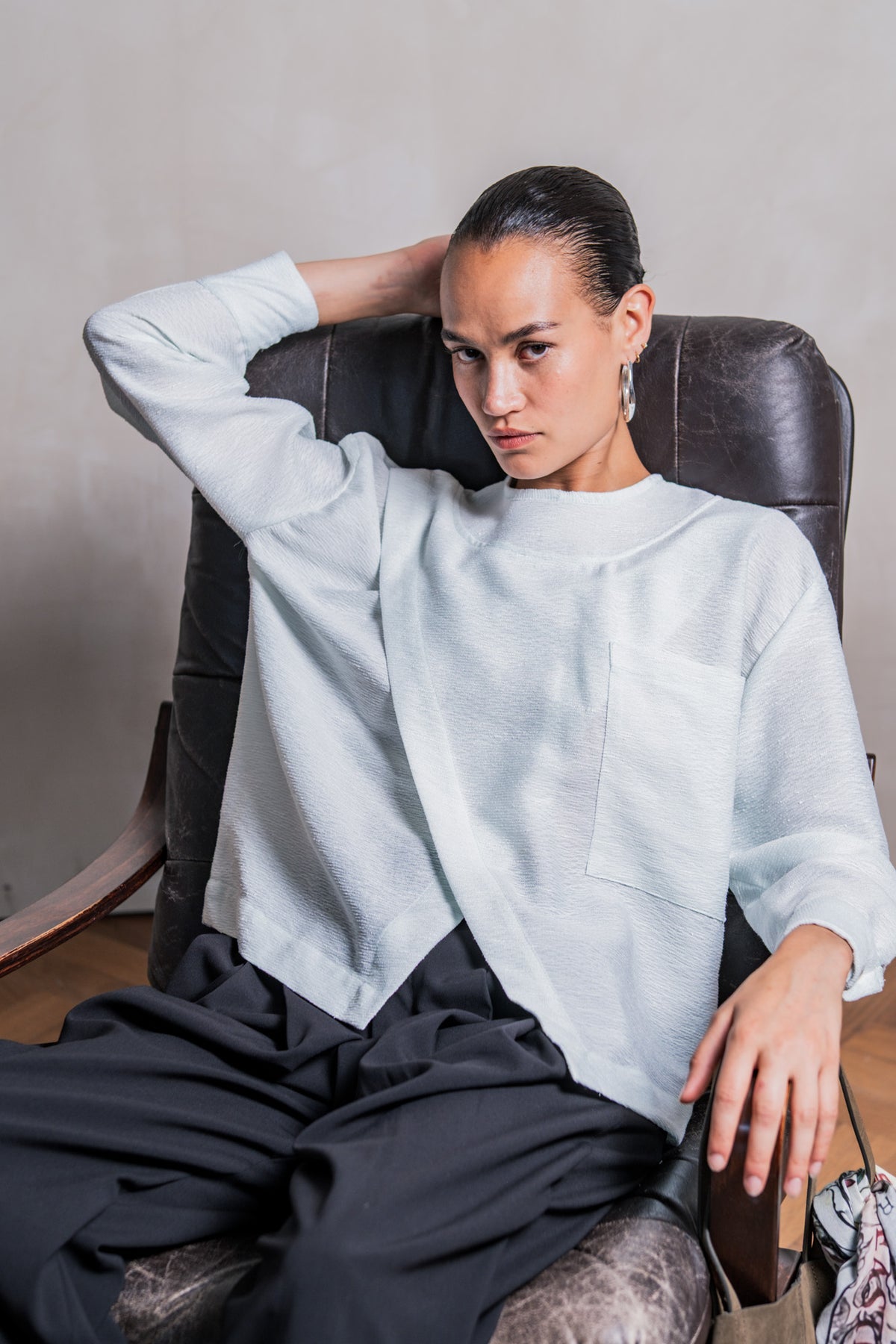 Woman wearing the Veya Blouse sewing pattern from Fibre Mood on The Fold Line. A top pattern made in crepe, viscose, poplin, lyocell, chambray, linen, seersucker, or broderie anglaise fabric, featuring a loose fit, round neck, asymmetrical overlap, pocket, and long sleeves with cuffs.
