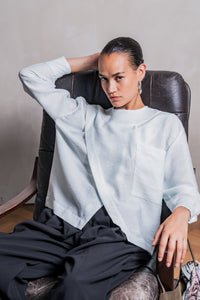 Woman wearing the Veya Blouse sewing pattern from Fibre Mood on The Fold Line. A top pattern made in crepe, viscose, poplin, lyocell, chambray, linen, seersucker, or broderie anglaise fabric, featuring a loose fit, round neck, asymmetrical overlap, pocket, and long sleeves with cuffs.