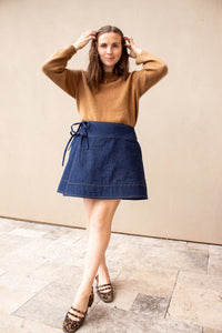 Woman wearing the Winnie Wrap Skirt sewing pattern from Madswick on The Fold Line. A skirt pattern made in cotton, linen, seersucker, denim, wool, twill, corduroy, or canvas fabric, featuring an A-line shape, ties and buckles, in-seam pockets, and mini length.