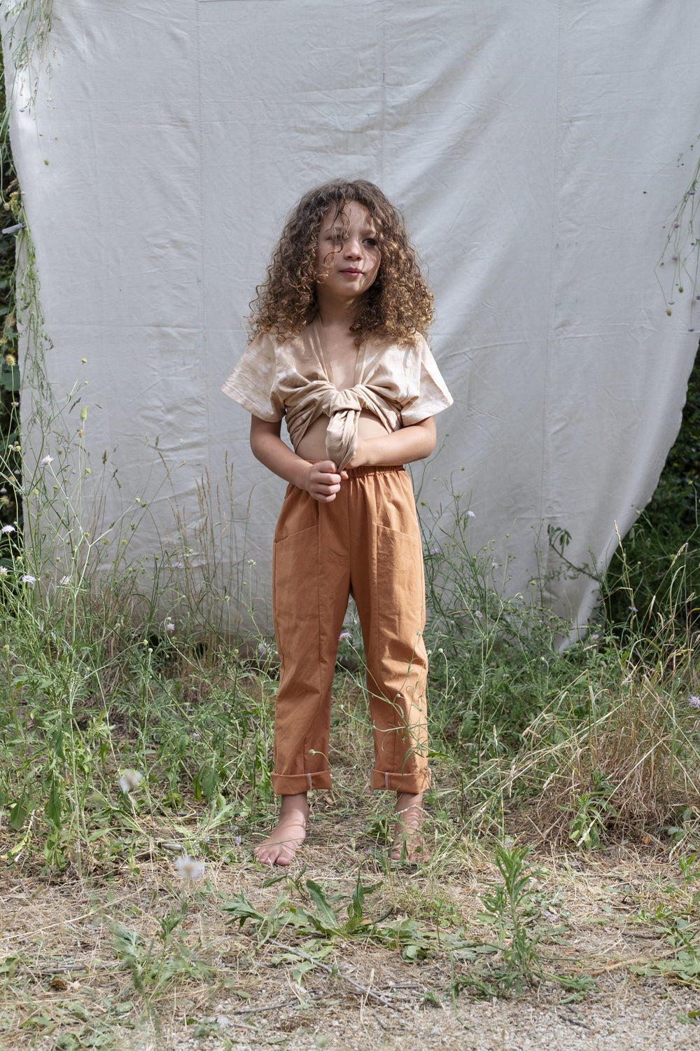 Child wearing the Pio Pants sewing pattern from Ready to Sew on The Fold Line. A trouser pattern made in cotton poplin, linen, batik, light denim or corduroy fabrics, featuring an elasticated waist, relaxed fit, two deep front pockets and two back patch p