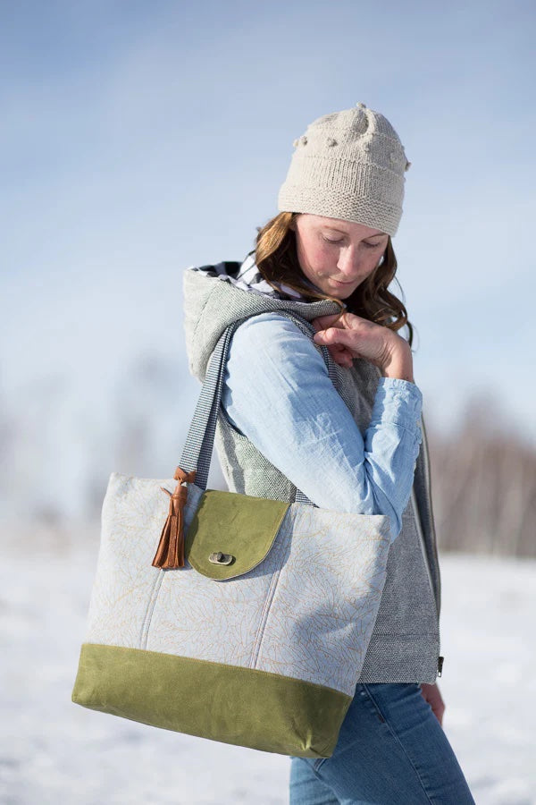 Woman holding the Explorer Tote sewing pattern from Noodlehead on The Fold Line. A tote pattern in made in waxed canvas, cotton canvas, cotton/linen canvas, denim or home dec fabrics, featuring a large size, front pocket, divided back pocket, large divide