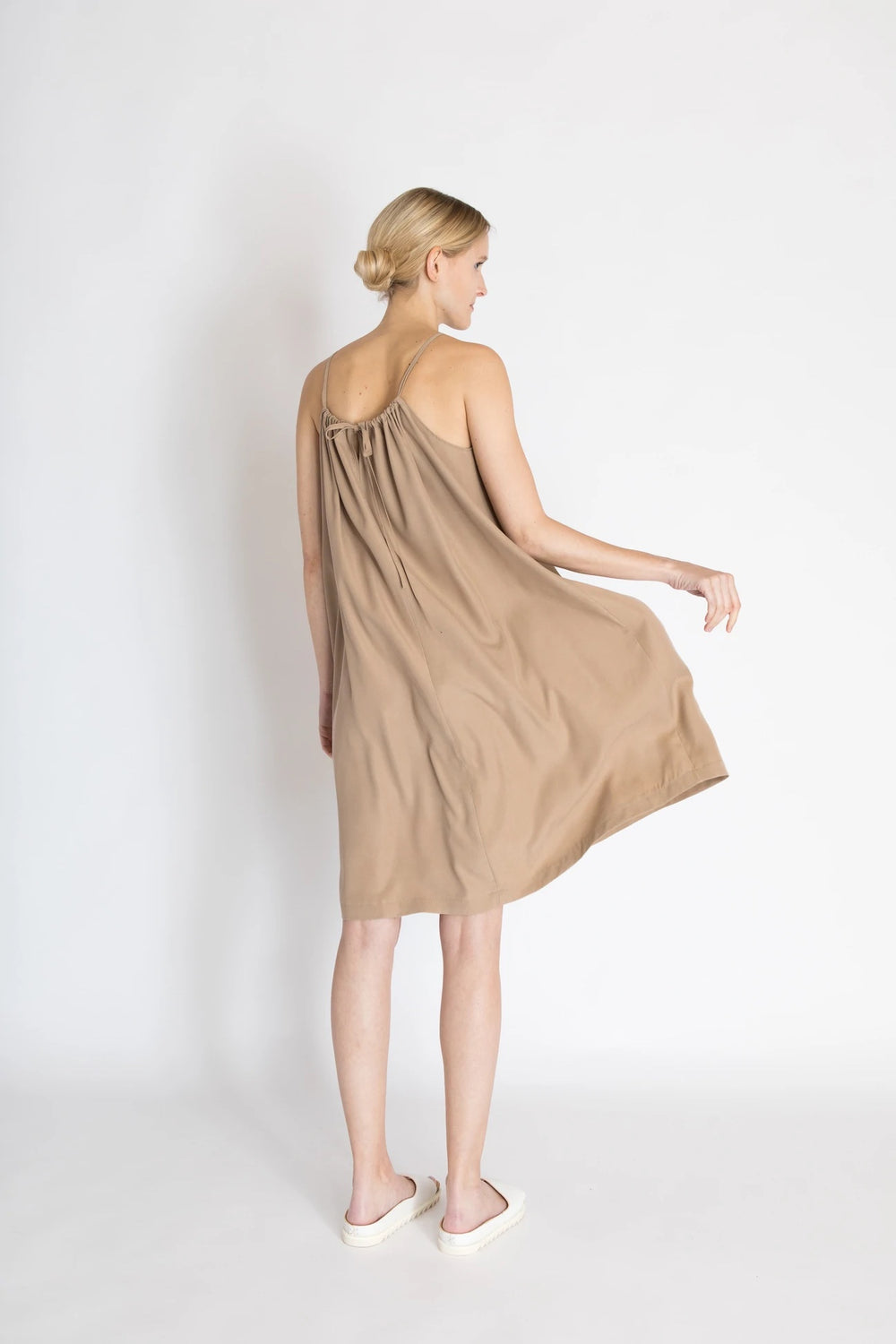 Woman wearing the Greta Dress sewing pattern from Bara Studio on The Fold Line. A sun dress pattern made in cotton, linen or tencel fabrics, featuring a loose wide fit, thin shoulder straps, side slits, neckline gathers, back neck bow closure and knee len