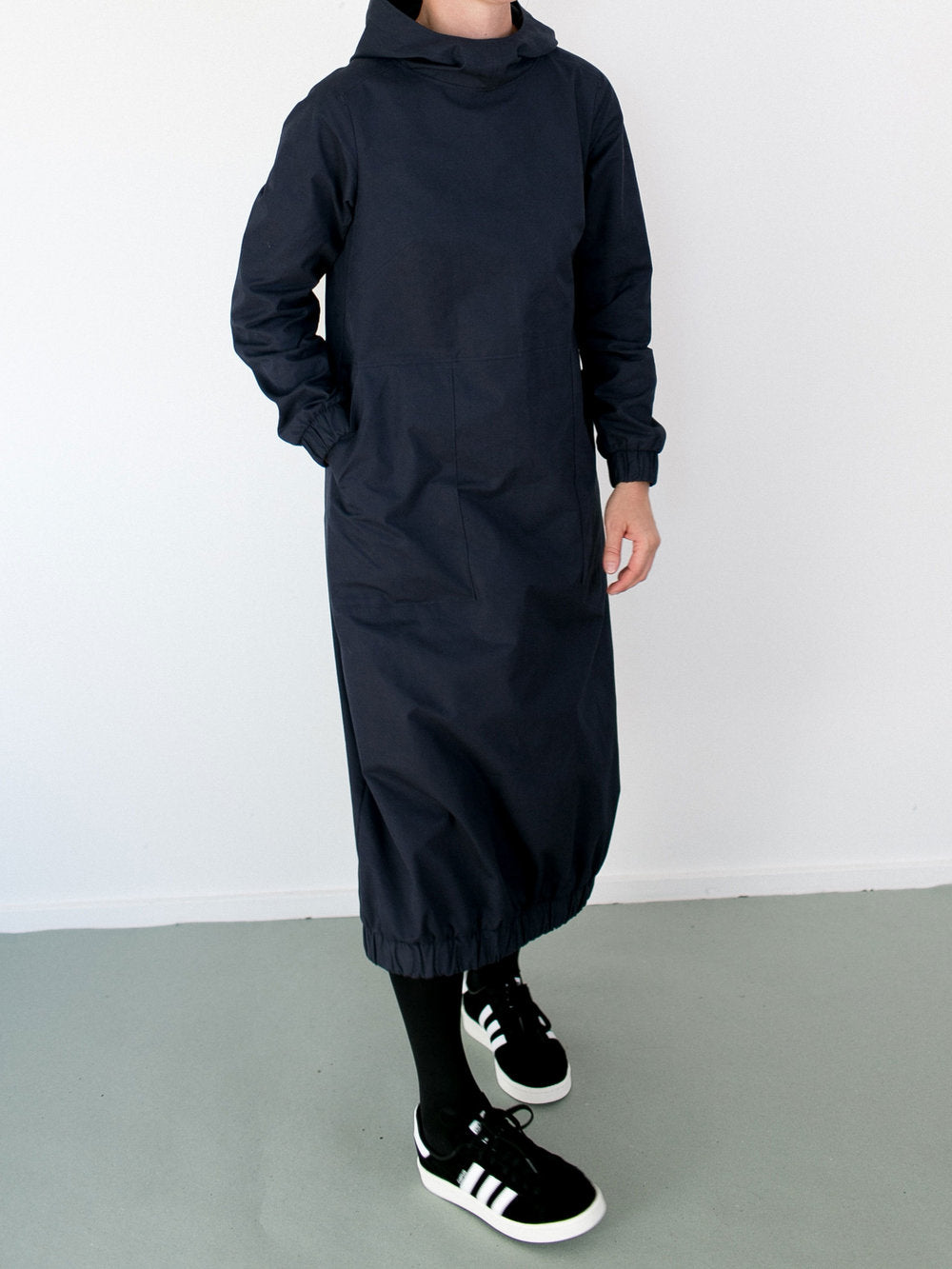 Woman wearing the Hoodie Dress sewing pattern by The Assembly Line. A relaxed fitting pullover dress pattern made in denim, linen or canvas fabric featuring elastic cuffs at sleeve and skirt bottom, a large front pocket and hood.