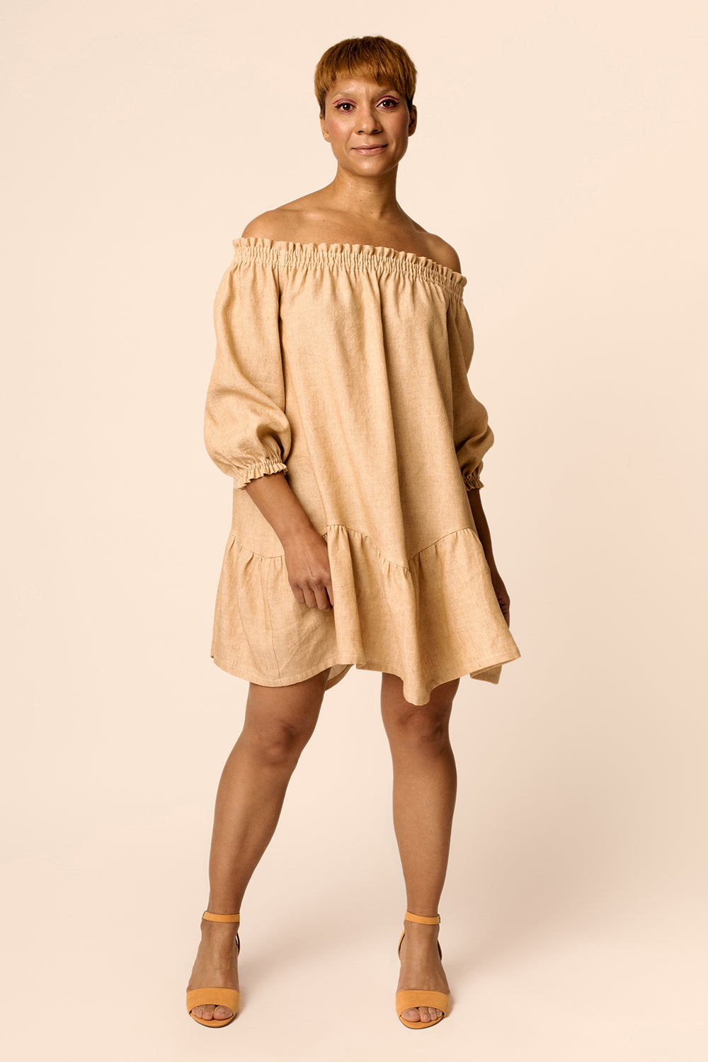 Woman wearing the Ilma Smock Dress sewing pattern from Named on The Fold Line. A dress pattern made in light linen, organic cotton, tencel or rayon fabrics, featuring an off-the-shoulder style, mini length, wavy tiered hemline, elasticated neckline, wrist
