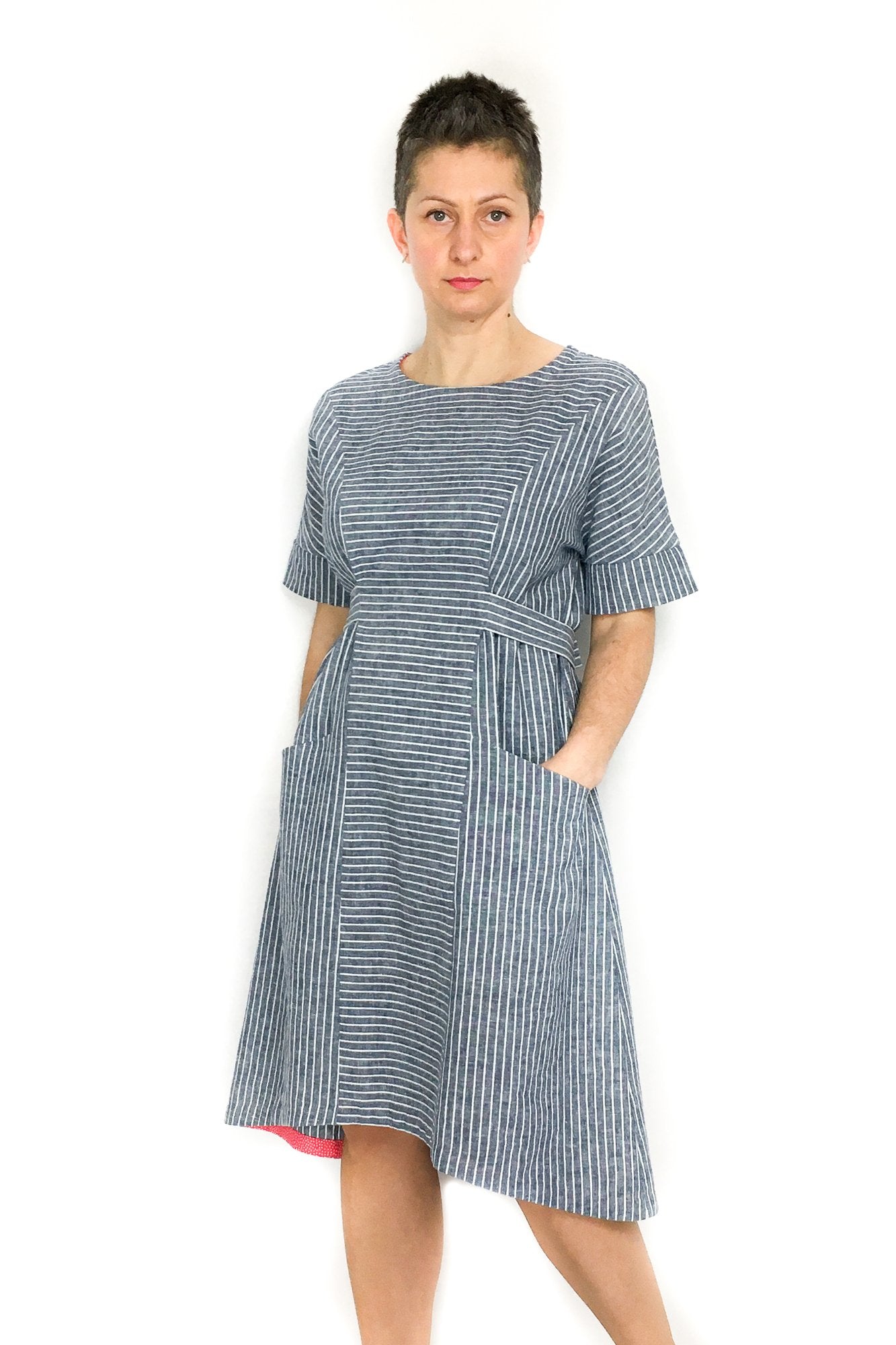 Woman wearing the Jasmine Dress sewing pattern by Dhurata Davies Patterns. A dress pattern made in any light/medium weight woven fabric with some drape, featuring a short sleeve with wide cuff, angled pockets, waist ties and a hem that is slightly raised 