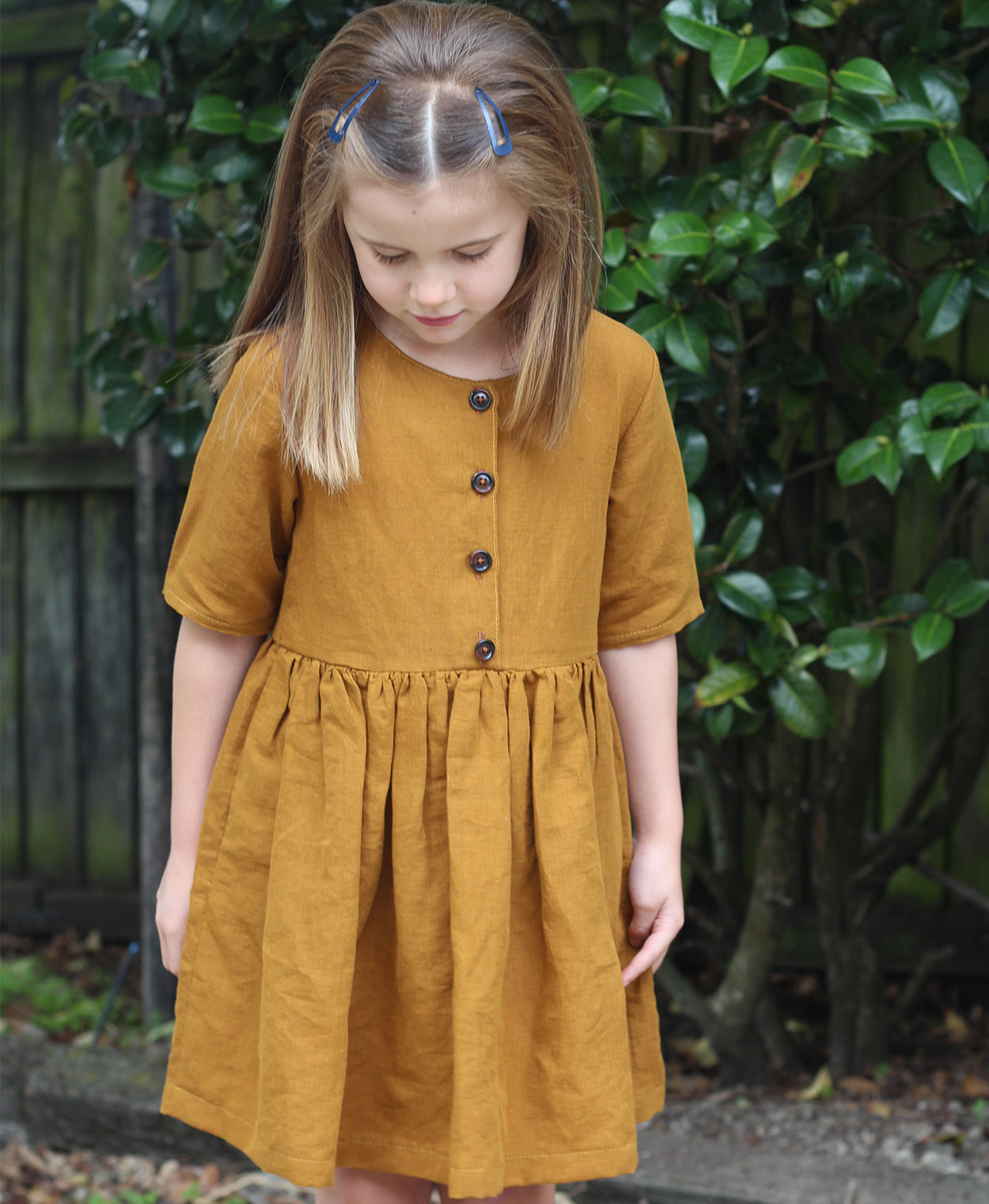 Child wearing the Baby/Child Kauri Dress sewing pattern by Below the Kowhai. A dress pattern made in light to medium weight linen, cotton, flannel or sateen fabrics, featuring a gathered waist, relaxed fit, pockets, elbow length sleeves and button bodice.