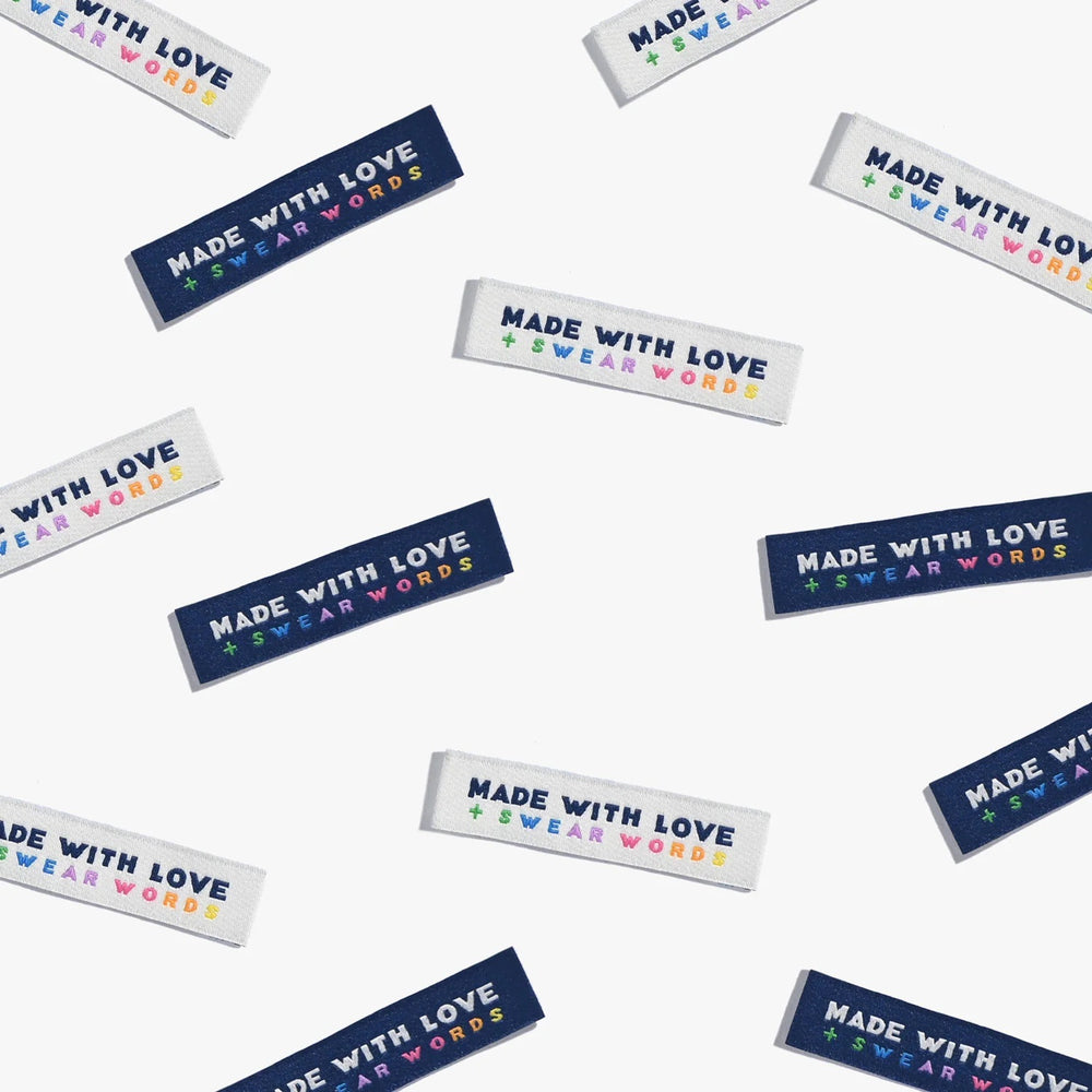 Photo showing 'Made With Love and Swear Words' Cotton Labels from Kylie & The Machine on The Fold Line. A washable, durable, and non-scratchy cotton label featuring a cream or navy background with bold navy or cream coloured text and rainbow mini text, th