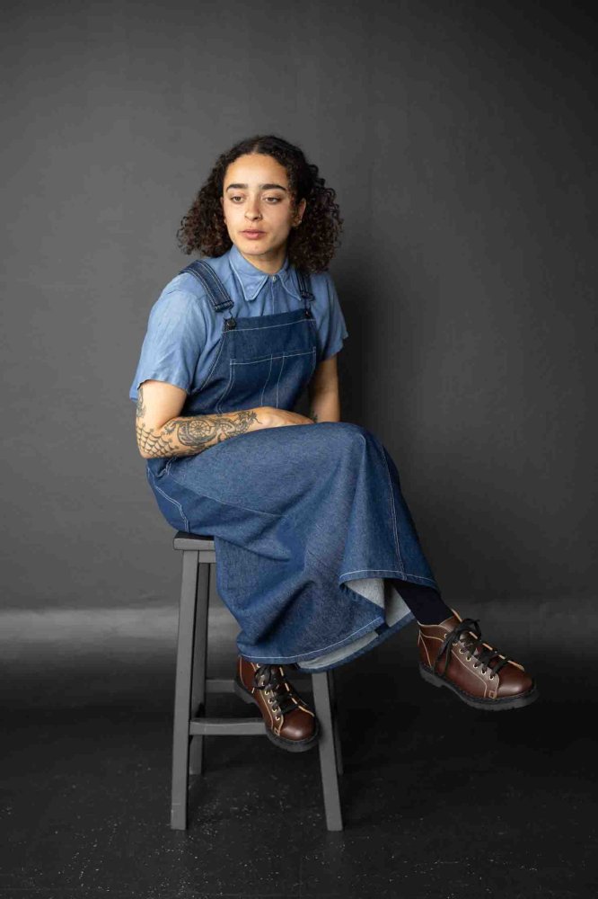 Woman wearing the Margo Dress sewing pattern from Merchant & Mills on The Fold Line. A pinafore dress pattern made in denim, cotton canvas, cotton drill, corduroy, and yarn-dyed medium weight linen fabrics, featuring topstitching, dungaree clips, midi len