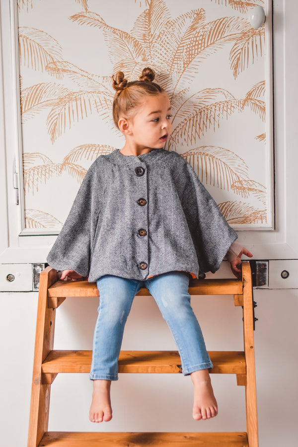 Child wearing the Mini Harry Cape sewing pattern from I AM Patterns on The Fold Line. A cape pattern made in boiled wool, wool gabardine, melton, bouclé or jacquard. fabrics, featuring a circular silhouette, in-seam pockets, flat collar, fully lined, fron