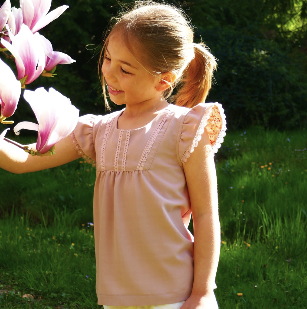 Child wearing the Child/Teen Obélie Blouse sewing pattern from Petits D’om on The Fold Line. A blouse pattern made in viscose, double gauze, Liberty, plumetis, cotton satin, batiste or tencel fabrics, featuring a front bib with gathers, rounded square nec