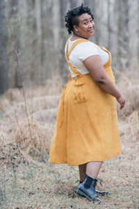 Woman wearing the Penny Pinafore sewing pattern by Sew Liberated. A pinafore pattern made in mid-weight woven fabrics, such as linen, gabardine, lightweight twill or denim, linen or silk noil fabrics, featuring a relaxed fit, roomy back pockets, and a dra