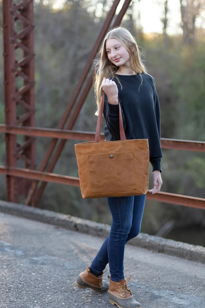 Woman holding the Pepin Tote sewing pattern from Noodlehead on The Fold Line. A tote pattern made in canvas or waxed canvas fabrics, featuring a front pocket with snap closure, divided interior pocket, recessed zip closure and shoulder length straps.