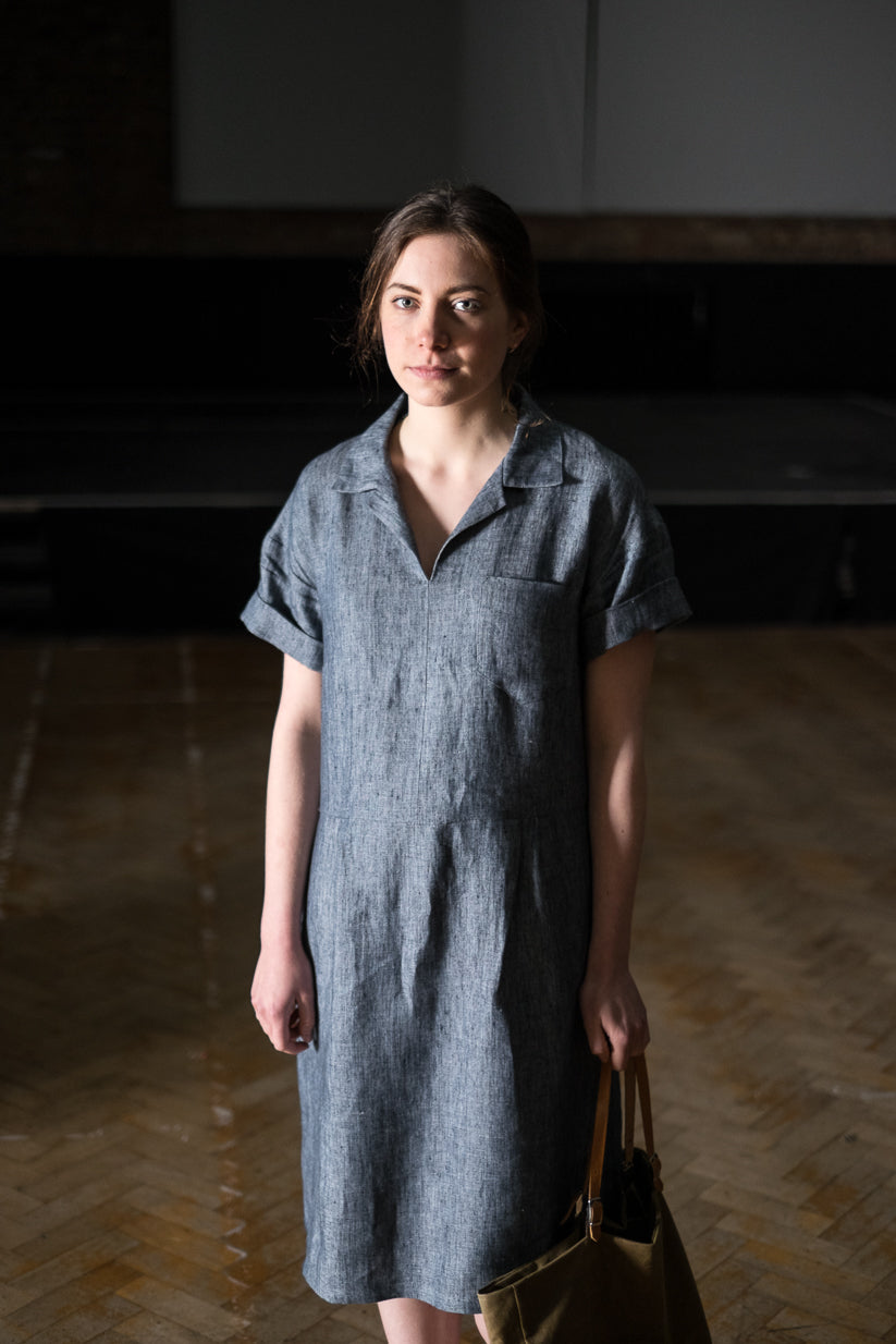 Woman wearing the Factory Dress sewing pattern by Merchant and Mills. A pull on dress pattern made in linen, cotton drill, lightweight denim, fine wool or corduroy fabric featuring rolled up short sleeves, breast pocket, collar with a V-neck and loose fit
