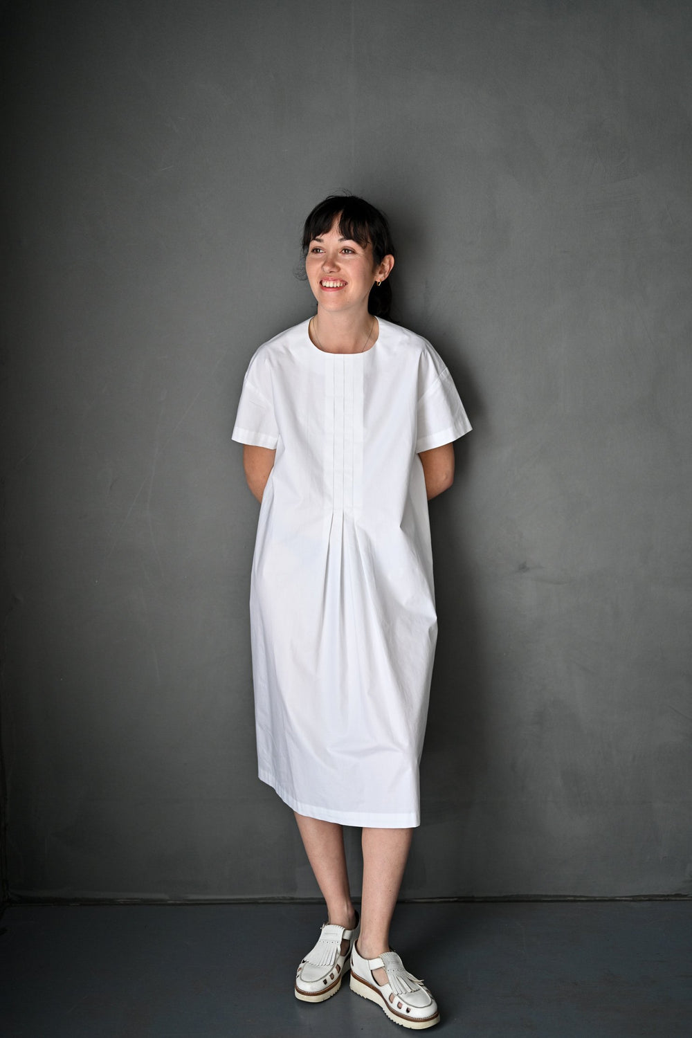 Woman wearing the Box Box Top and Dress sewing pattern by Merchant and Mills. A boxy dress pattern made in poplin, chambray, denims or lightweight wool fabric featuring a front pleat detail, rouleaux loop back neck closure, round neck and short sleeves.