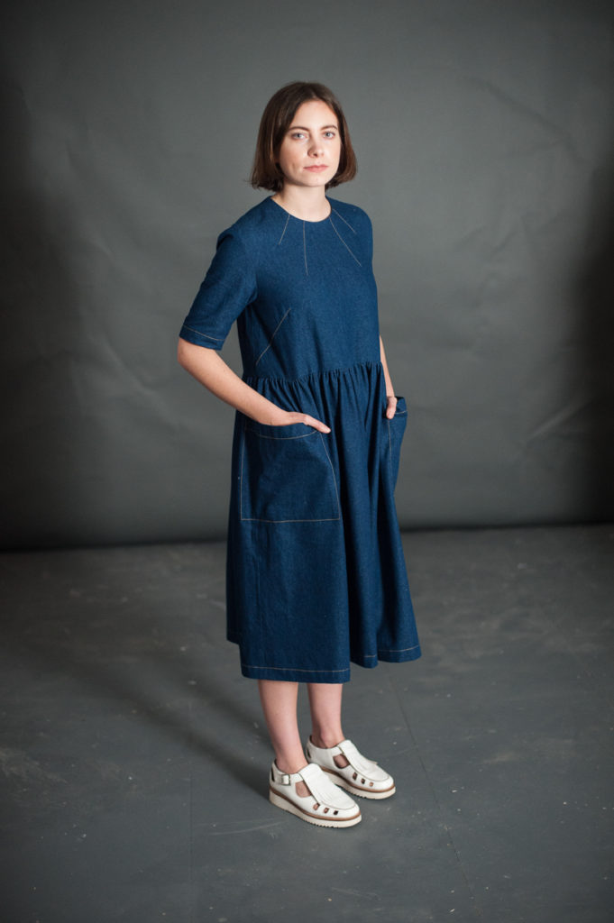 Woman wearing the Ellis Dress sewing pattern by Merchant and Mills. A dress pattern made in linen, light wools, and light to medium weight cotton fabric featuring four delicate neck darts, set in elbow length sleeves, gathered waist and bold patch pockets