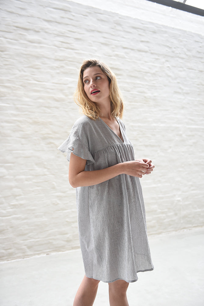 Woman wearing the Rosalie Dress sewing pattern from Fibre Mood on The Fold Line. A dress pattern made in cotton, poplin or linen blend fabrics, featuring a babydoll style, deep V-neck, short ruffled sleeves, roomy fit, above knee length, front and back yo