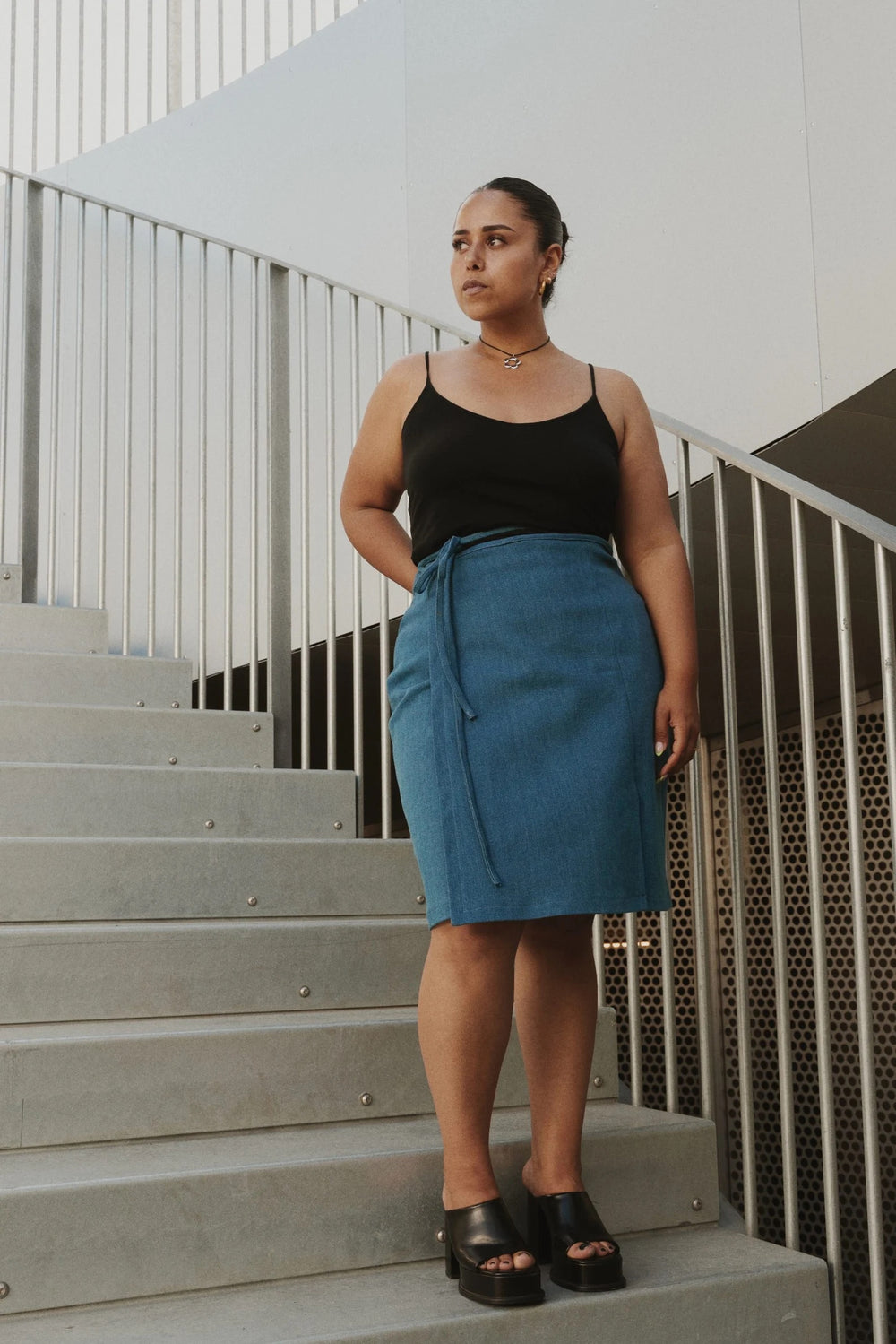 Woman wearing the Strap Wrap Skirt sewing pattern from Puff and Pencil on The Fold Line. A wrap skirt pattern made in light jacquard, satin, thin wool qualities, linen and light denim fabrics, featuring a high-waist, knee-length, waist ties, front left si