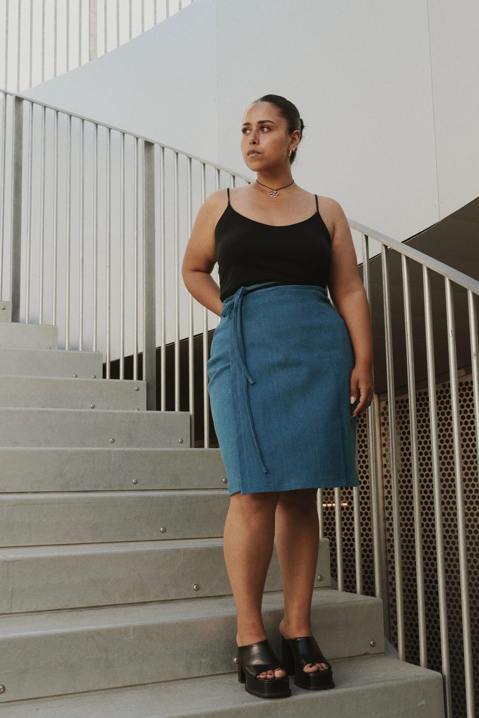 Woman wearing the Strap Wrap Skirt sewing pattern from Puff and Pencil on The Fold Line. A wrap skirt pattern made in light jacquard, satin, thin wool qualities, linen and light denim fabrics, featuring a high-waist, knee-length, waist ties, front left si