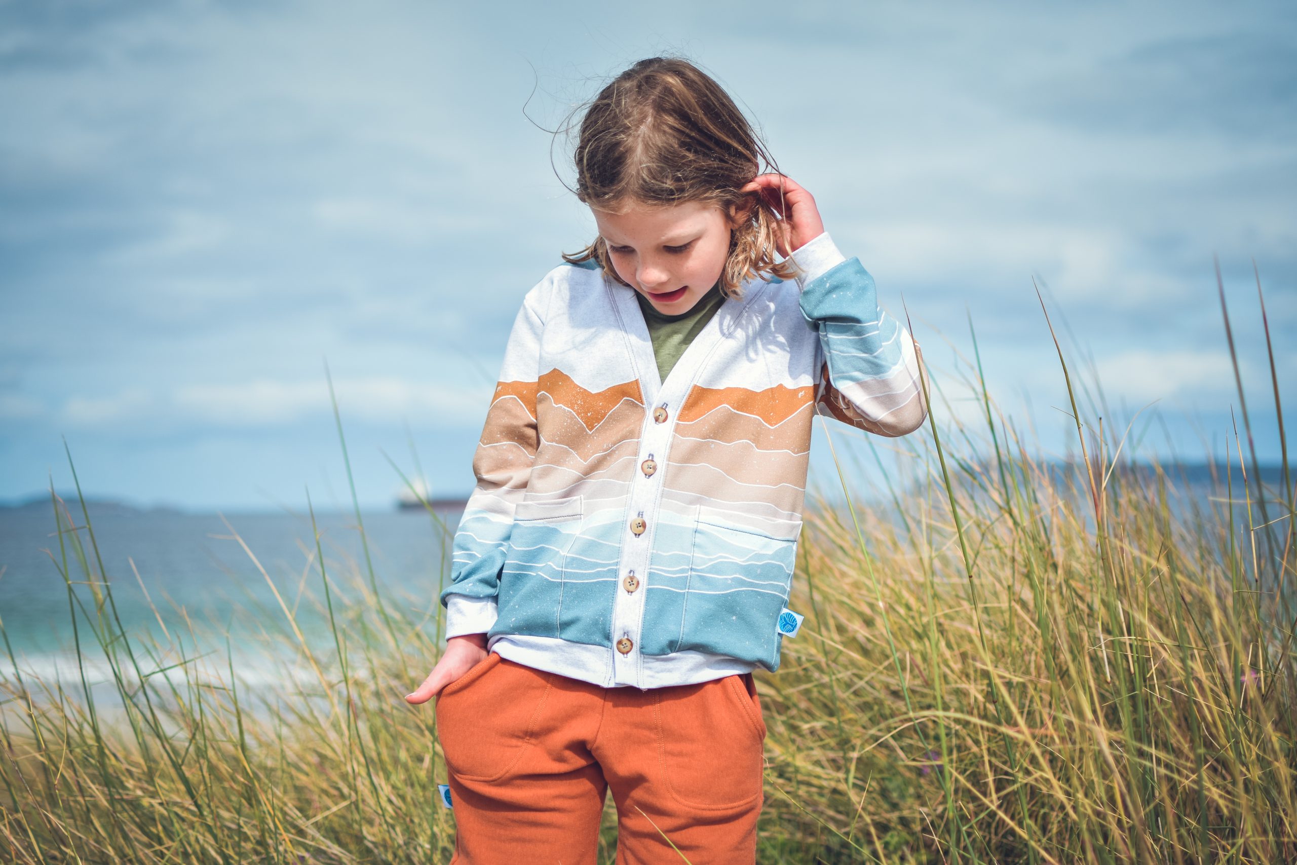 Child wearing the Baby/Child Salt Air Cardigan sewing pattern from Waves & Wild on The Fold Line. A cardigan pattern made in cotton/Lycra, french terry or sweatshirt fleece fabrics, featuring a slim fit, patch pockets, V-neck, button front closure and ful