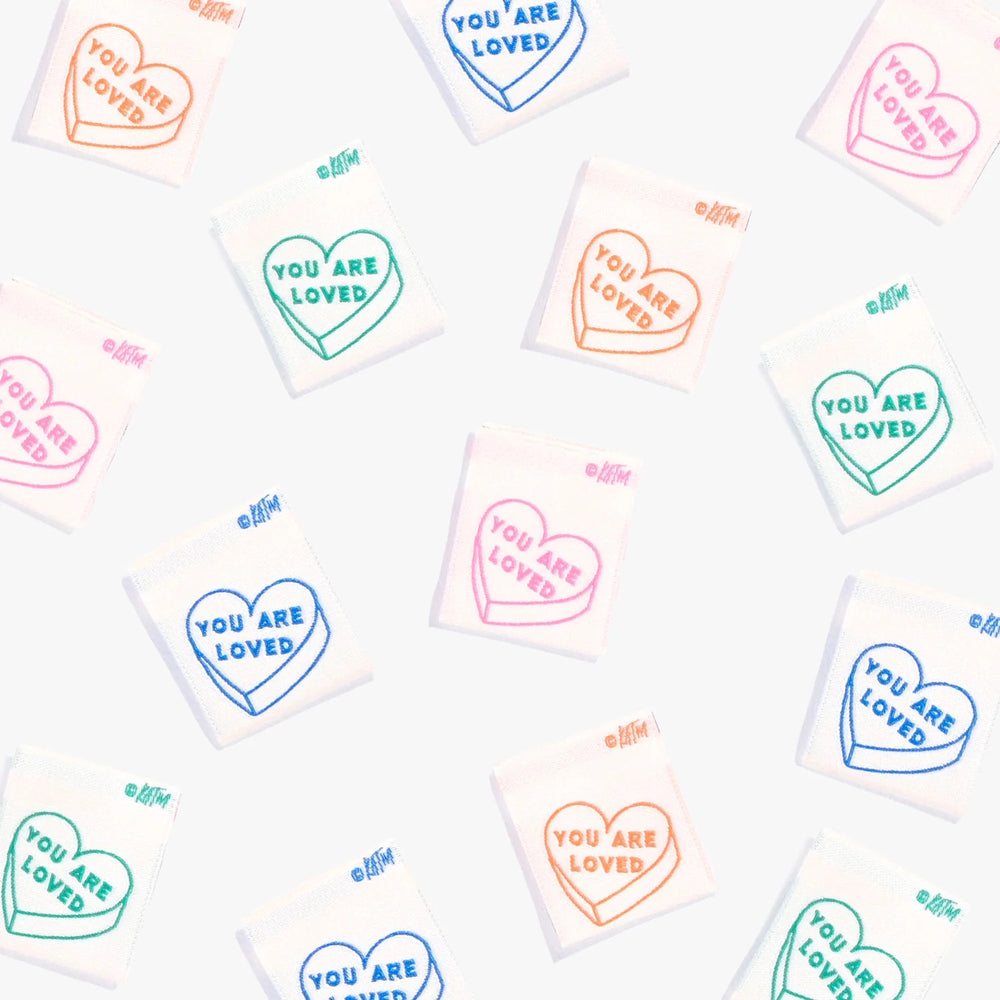 Photo showing 'You Are Loved’ Labels from Kylie & The Machine on The Fold Line. A washable, durable, and non-scratchy label. Included are 6 double-sided woven labels with text in orange, mid-pink, mid-green or royal blue on a lush cream background. They a