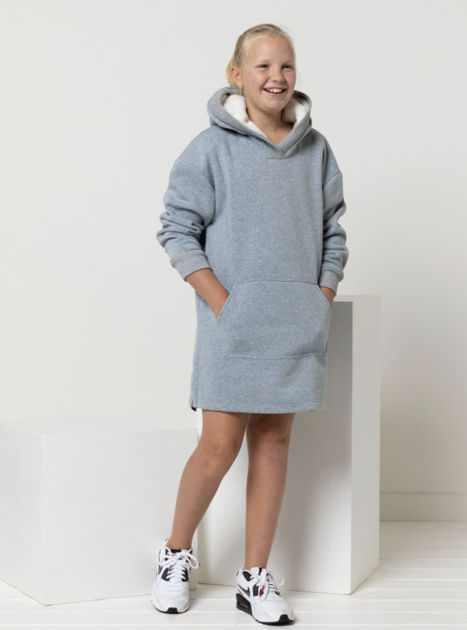 Child wearing the Child/Teen Zara Hooded Dress sewing pattern from Style Arc on The Fold Line. A knit dress pattern made in Sweater knit or French terry fabrics, featuring a lined hood, sleeves with cuffs, centre front pocket, and side splits finished wit