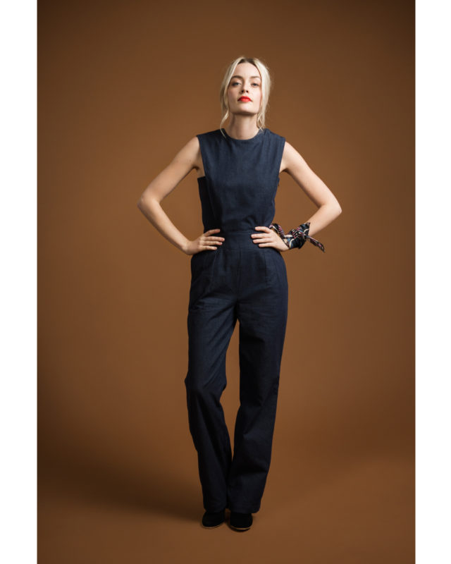 Woman wearing the Acacia Jumpsuit sewing pattern from Orageuse on The Fold Line. A sleeveless jumpsuit pattern made in canvas, light denim or serge fabrics, featuring a bib with slanted pleats from shoulder to waist at the front and back, crew neck, inver